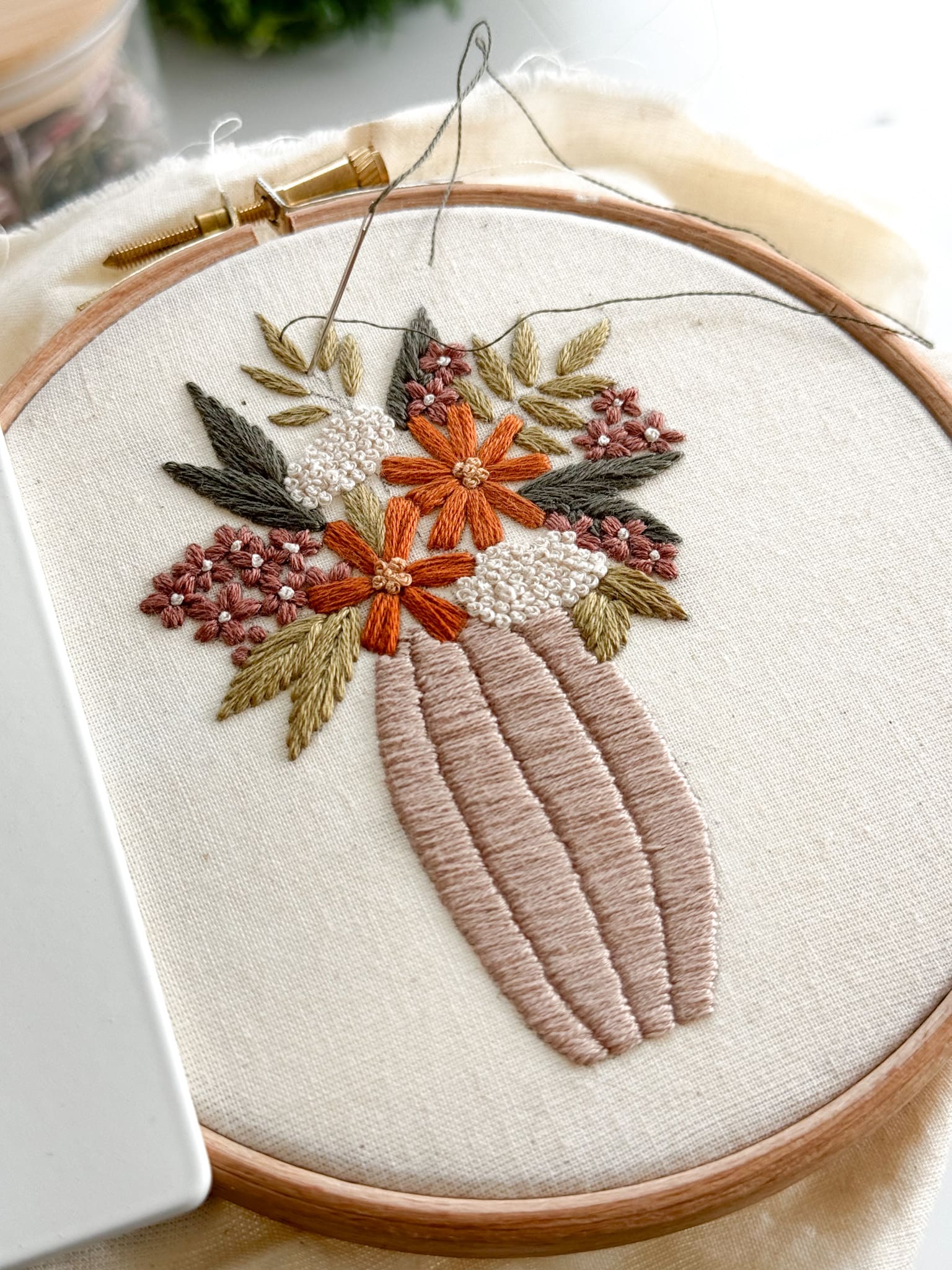 An example of a beginner-friendly embroidery pattern that is included in embroidery membership, Bloom Embroidery Academy. An embroidery hoop filled with cotton calico fabric, stitched with a beige vase filled with orange and dusky pink flowers, and surrounded by warm green leaves.