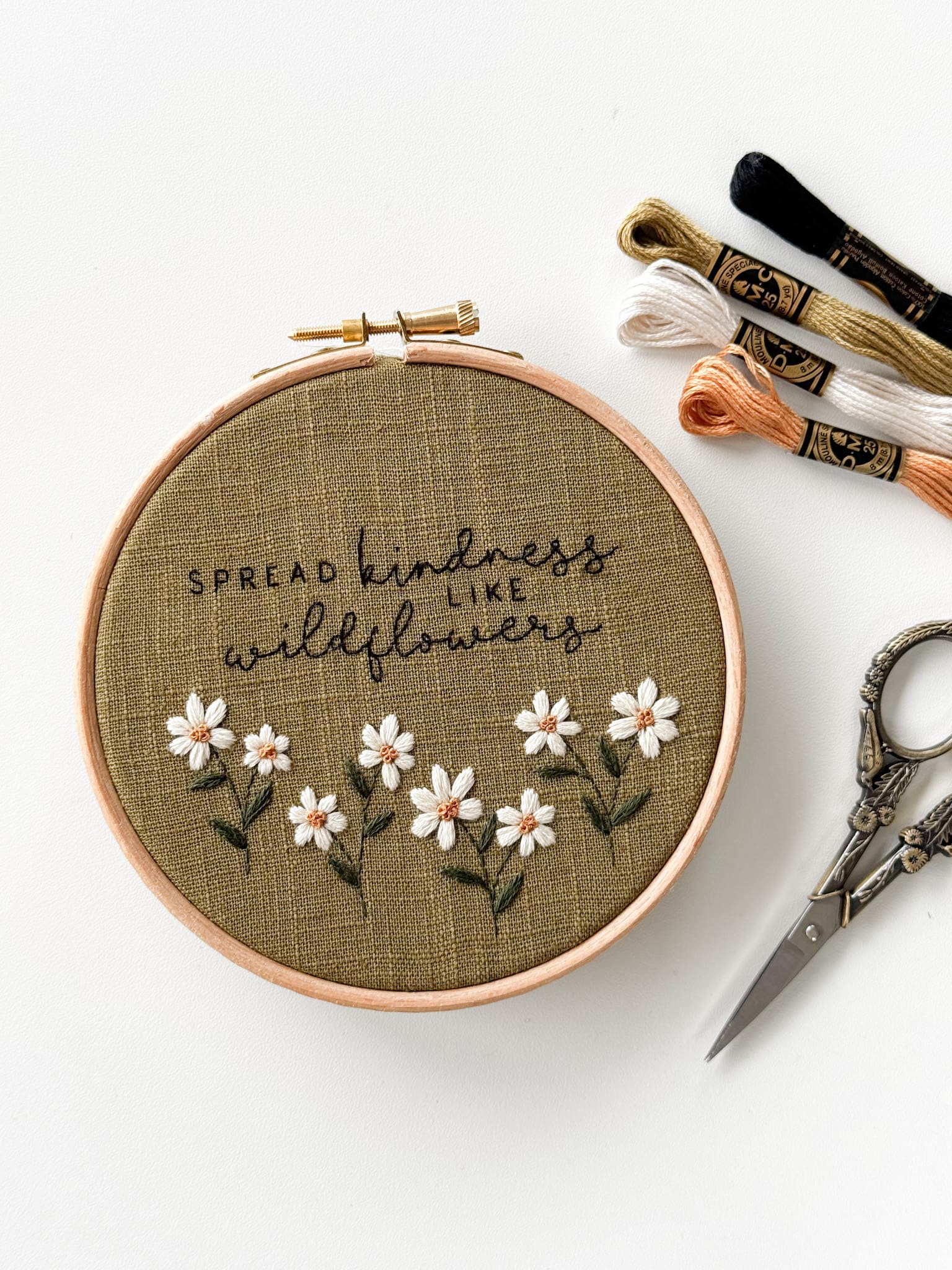 An example of a beginner-friendly embroidery pattern that is included in embroidery membership, Bloom Embroidery Academy. An embroidery hoop filled with green linen fabric with the quote "spread kindness like wildflowers", with daisies lined out below.