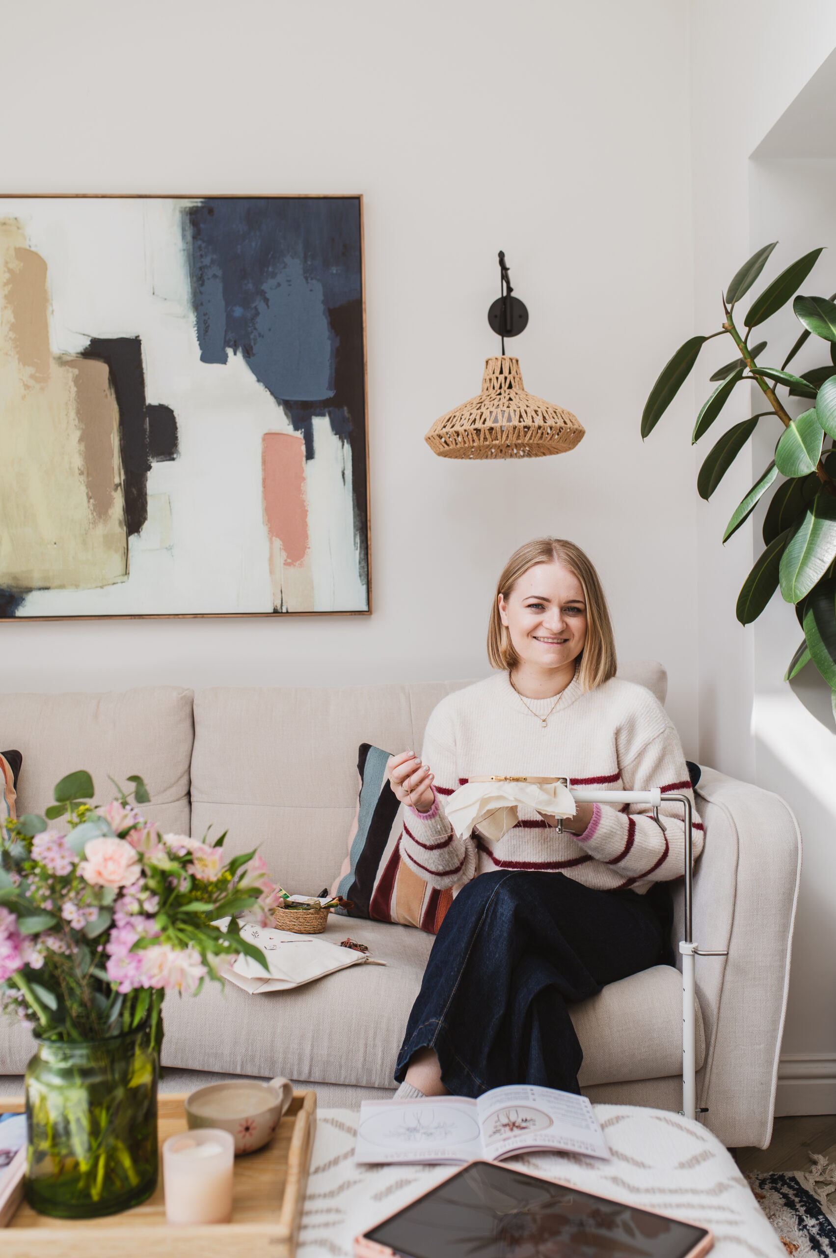 Sophie sat on the sofa with her embroidery hoop in her stand, holding her working thread as she stitches and smiles at the camera