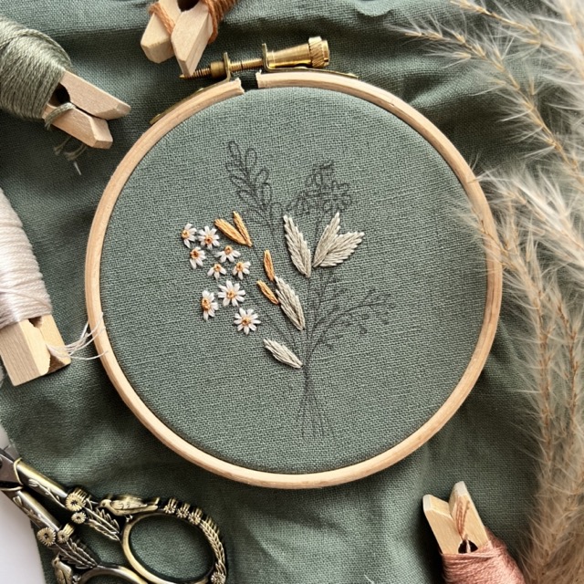flatlay of the 'summer harvest' embroidery pattern on green fabric, surrounded by scissors and thread wound on pegs